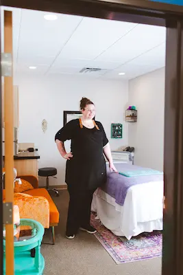 Kelli smiling and standing beside a prepared massage table, waiting for a client.